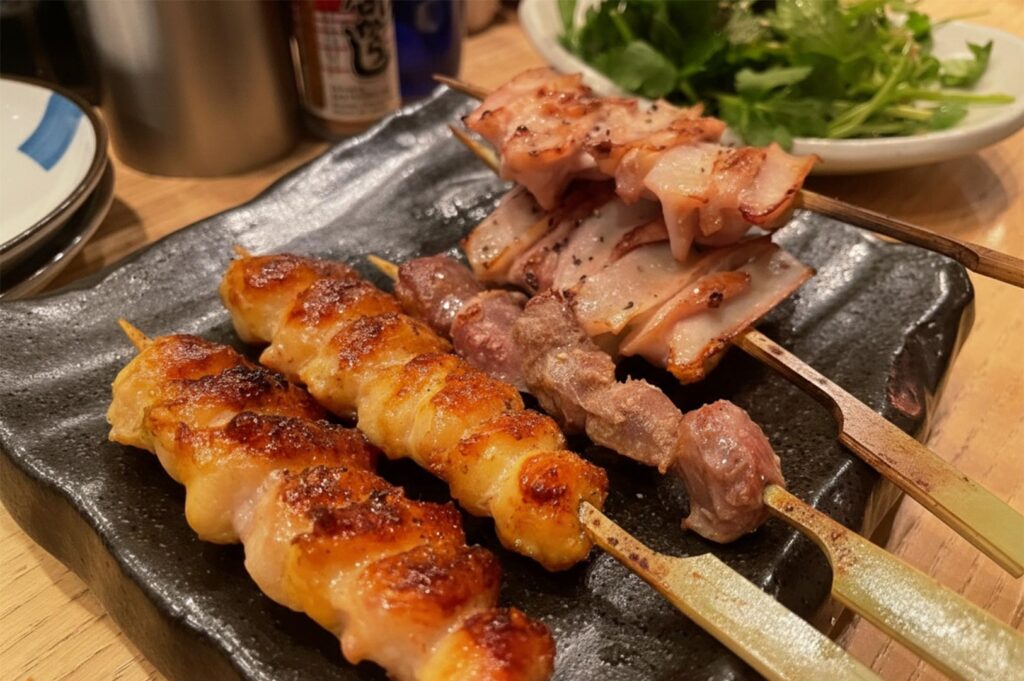 A close-up shot of various grilled chicken skewers, including chicken skin and thigh, served on a textured black ceramic plate in a restaurant.