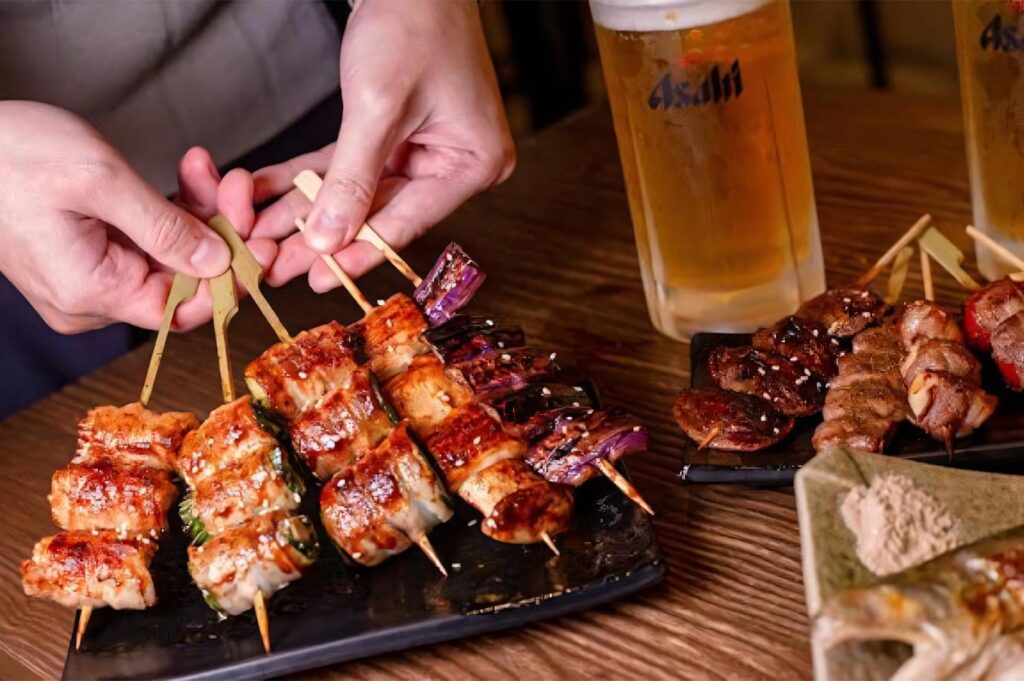 A close-up of various grilled yakitori skewers being served on a wooden table next to a cold mug of Asahi draft beer.