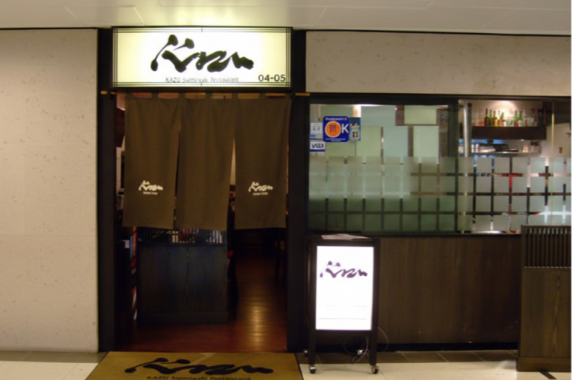 The exterior entrance of Kazu Sumiyaki Restaurant, featuring a dark brown noren curtain, a backlit sign with Japanese calligraphy, and glass windows.
