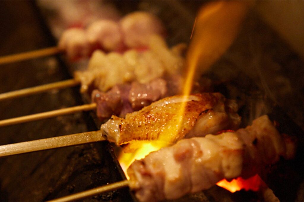 An action shot of chicken skewers being grilled over glowing binchotan charcoal with visible flames, capturing the traditional Japanese grilling process.