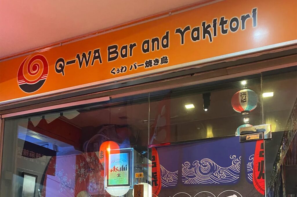 A close-up shot of the orange Q-WA Bar and Yakitori storefront sign with Japanese text and an illuminated Asahi beer neon sign in the window.