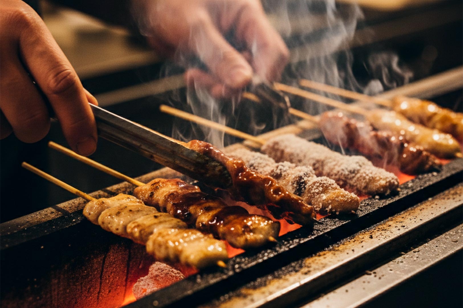 Close-up of a chef using tongs to grill various chicken yakitori skewers over a steaming charcoal robata grill.