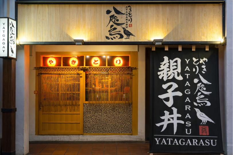 The storefront of "Yatagarasu" yakitori restaurant at night, featuring a wooden facade, glowing lanterns, and a large black banner with bold Japanese calligraphy.