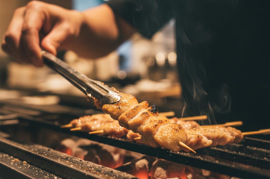 Close-up of a chef’s hand using metal tongs to flip a chicken skewer over a traditional Japanese grill.