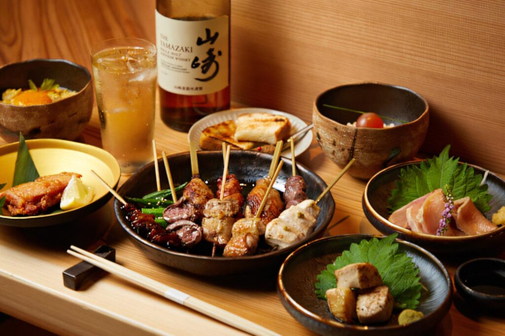 A complete Japanese dinner spread featuring assorted yakitori skewers, small side dishes, chicken sashimi, and a bottle of Yamazaki single malt whiskey.