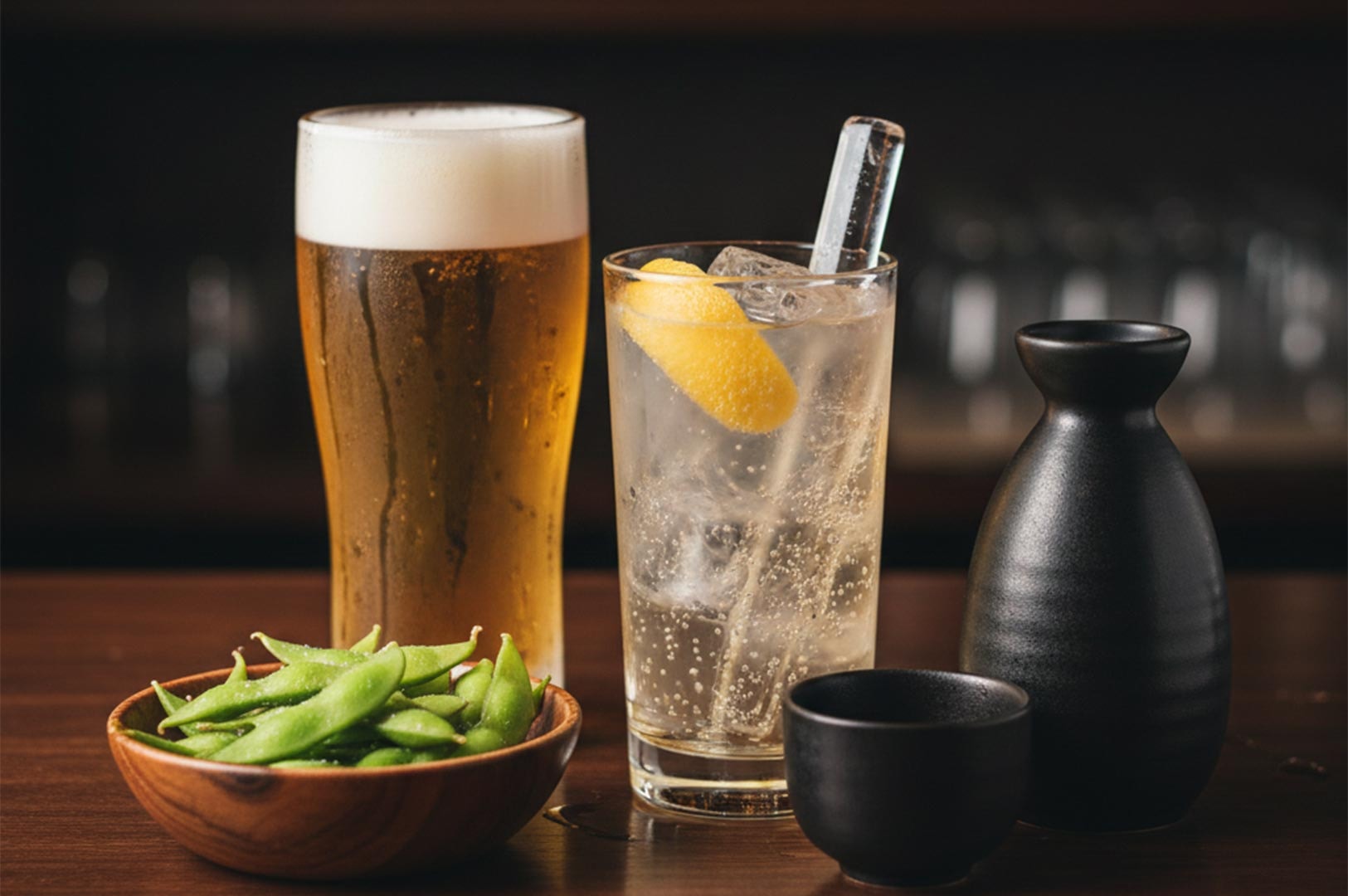A close-up of a Japanese drink set featuring a tall glass of beer, a lemon highball, a ceramic sake carafe, and a bowl of salted edamame.