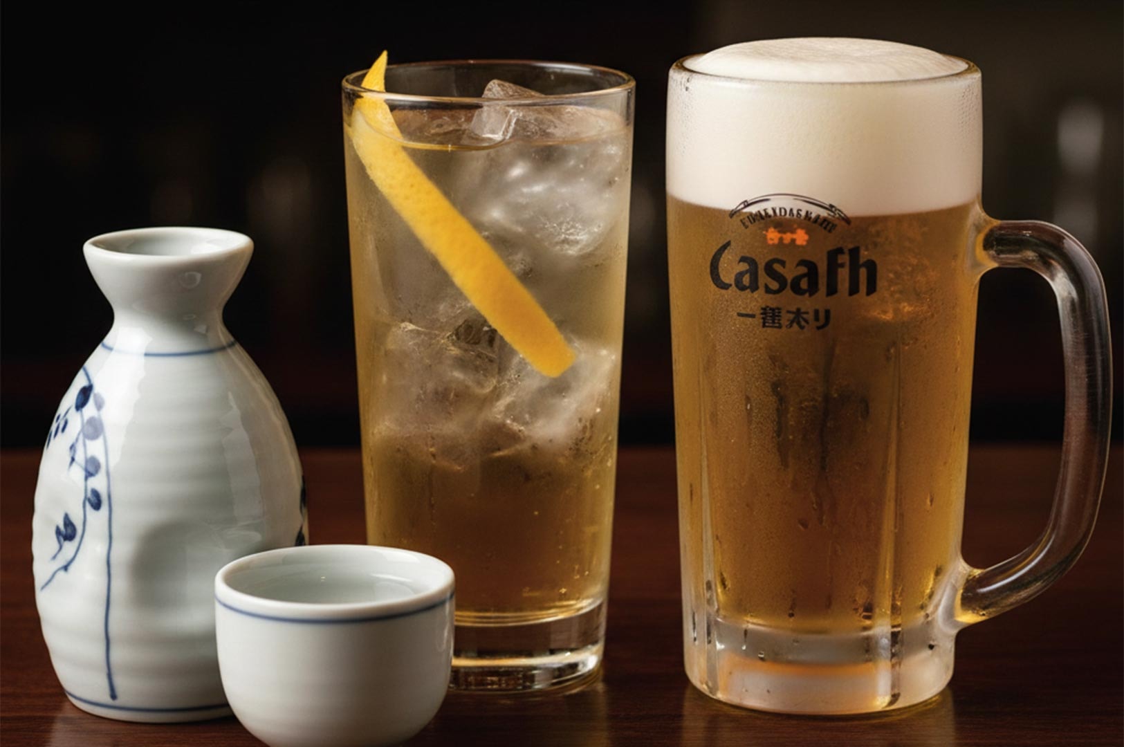 A selection of Japanese bar drinks featuring a ceramic sake carafe, a whiskey highball with lemon, and a frothy mug of draft beer.