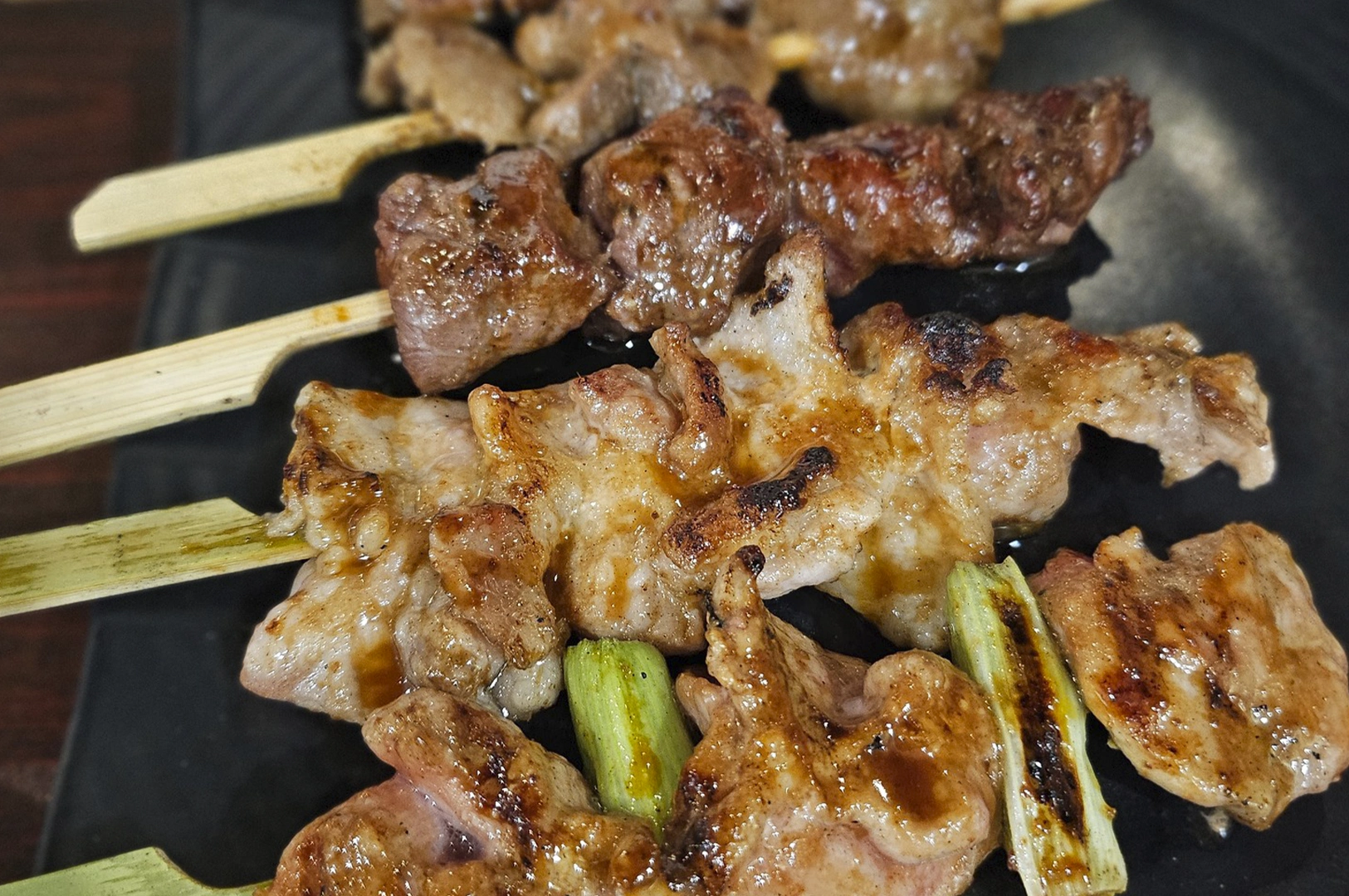 A close-up, high-angle shot of several grilled meat yakitori skewers resting on a dark, textured plate, showing charred, glazed textures and pieces of green scallion between the meat.