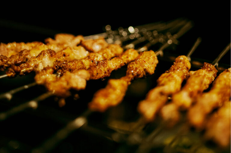 Close-up of several chicken yakitori skewers being grilled over a dark, glowing heat source.