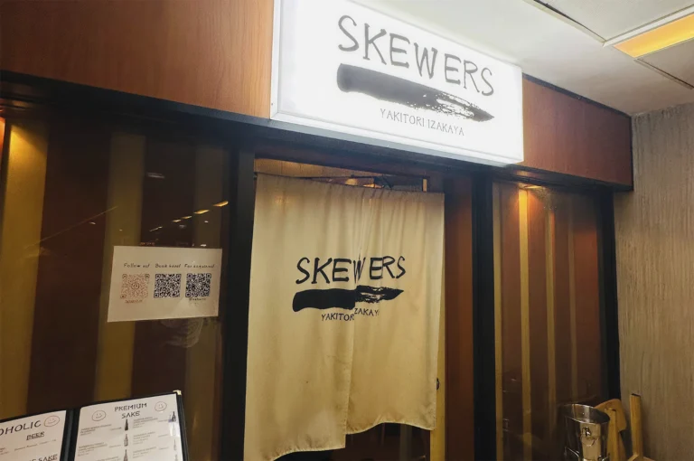 A medium shot of the storefront for "Skewers Yakitori Izakaya." Above the entrance is a glowing white rectangular sign featuring the word "SKEWERS" in a rustic, hand-drawn font above a bold black brushstroke. A cream-colored fabric noren curtain hangs in the doorway, repeating the logo. To the left, a glass window displays a white flyer with three QR codes for social media and bookings, and a menu board at the bottom left shows sections for "Premium Sake" and "Beer." The storefront has dark wood framing and warm, dim interior lighting.