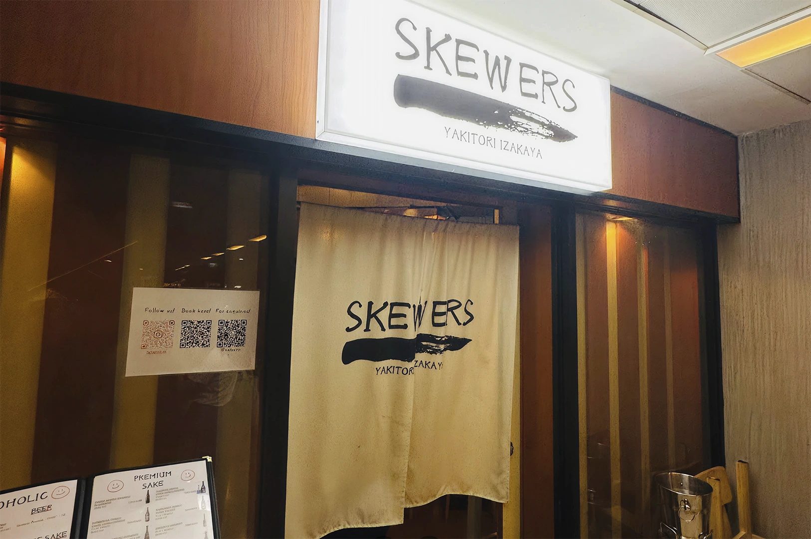 A medium shot of the storefront for "Skewers Yakitori Izakaya." Above the entrance is a glowing white rectangular sign featuring the word "SKEWERS" in a rustic, hand-drawn font above a bold black brushstroke. A cream-colored fabric noren curtain hangs in the doorway, repeating the logo. To the left, a glass window displays a white flyer with three QR codes for social media and bookings, and a menu board at the bottom left shows sections for "Premium Sake" and "Beer." The storefront has dark wood framing and warm, dim interior lighting.