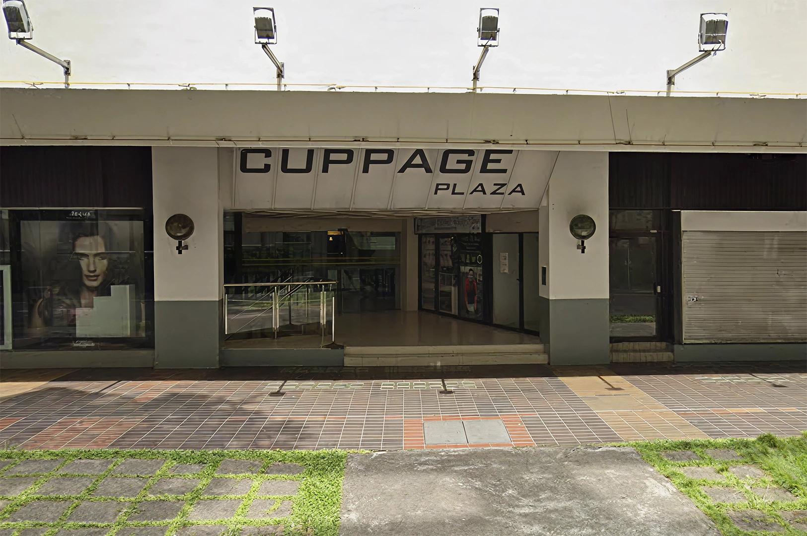 An exterior view of the Cuppage Plaza entrance, featuring a white sign with black text, a dark-tinted storefront, and outdoor lighting fixtures mounted above the signage.