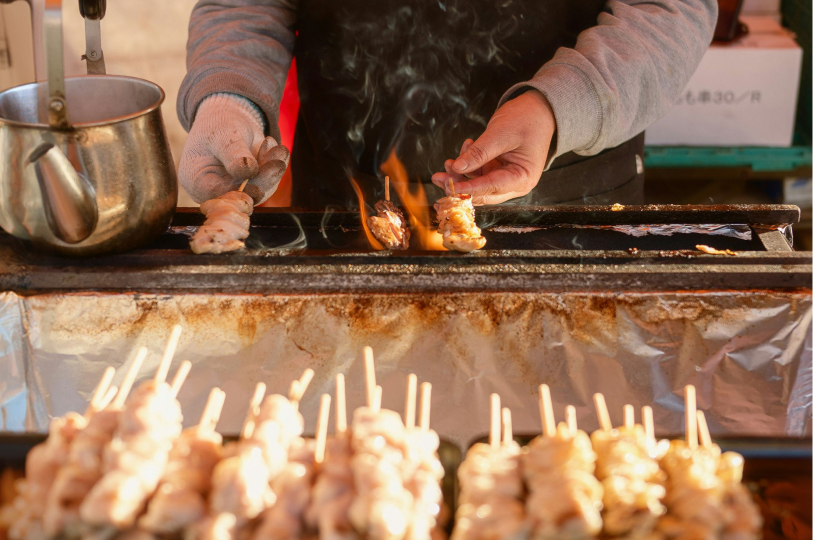 A street food vendor grilling chicken skewers over an open flame on a narrow charcoal grill.
