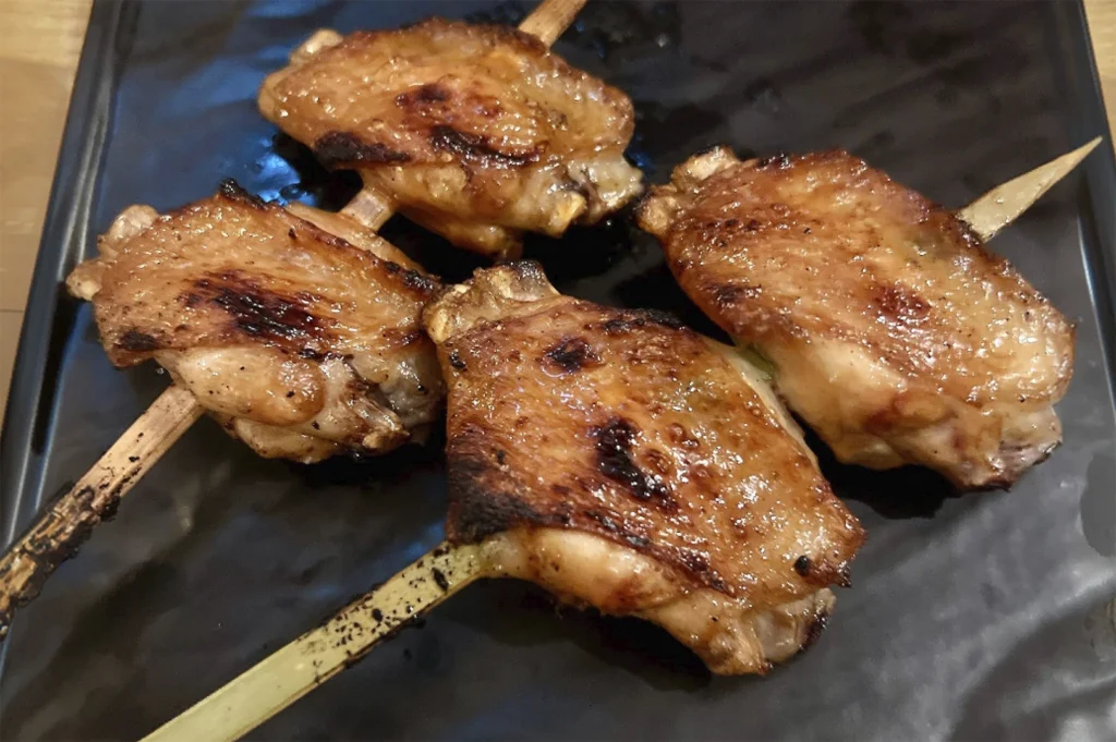 A close-up, top-down view of four grilled yakitori chicken wings served on a textured matte black plate. Each wing is skewered on a flat bamboo stick and features golden-brown, crispy skin with visible charred spots from grilling. The chicken has a glossy sheen, suggesting a light glaze or natural oils. The lighting is warm, highlighting the texture of the seared skin and the slightly burnt edges of the wooden skewers.
