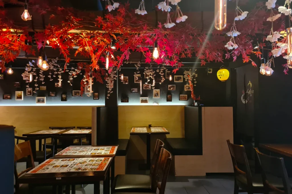 The interior of a dimly lit, atmospheric Japanese restaurant featuring an elaborate ceiling installation of red autumn maple leaves and delicate white cherry blossoms. Warm, vintage-style Edison bulbs and a single yellow paper lantern hang among the foliage, casting a soft glow over dark wooden tables and booths. The back wall is decorated with a series of small, framed photographs and a glowing light strip that highlights the clean lines of the modern furniture.