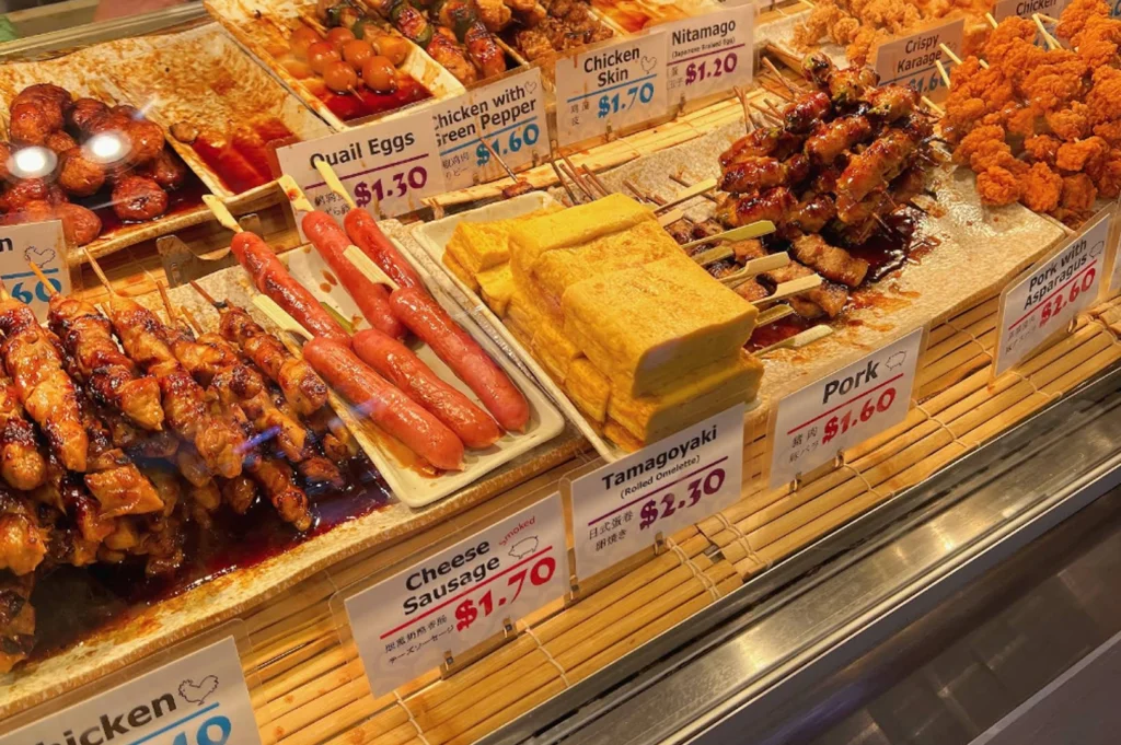 a glass display case filled with various Japanese yakitori skewers. The skewers are arranged on rectangular white platters resting on a bamboo mat. Options include Chicken Skin, Quail Eggs, Tamagoyaki (rolled omelette), Cheese Sausage, Pork, and Crispy Karaage. Each item is labeled with a small white sign showing the name in English and Japanese, along with prices ranging from $1.20 to $2.60. The grilled meats are coated in a glossy, dark brown tare sauce, reflecting the bright overhead lights of the display.