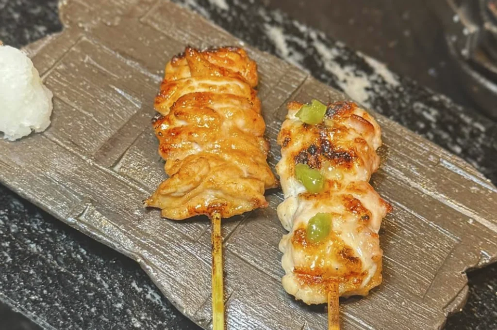 A close-up, high-angle shot of two yakitori skewers resting on a textured, dark gray ceramic plate. The skewer on the left features grilled chicken skin or thigh with a glistening, golden-brown glaze. The skewer on the right consists of succulent chicken pieces topped with small dollops of bright green wasabi paste. A small mound of grated daikon radish sits on the side of the plate against a dark, speckled background.
