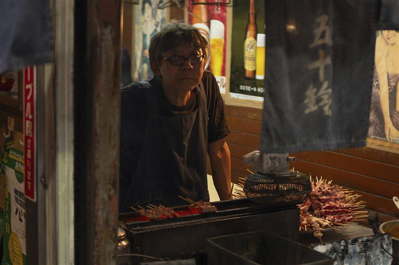 A candid, atmospheric shot of an elderly Japanese chef wearing glasses and a dark blue apron, standing behind a narrow charcoal grill. He has a neutral, focused expression. In the foreground, several skewers are cooking over the heat, while a small electric fan is positioned to blow smoke away. The background is warm and dimly lit, adorned with vintage Japanese beer posters and a dark "noren" curtain with white kanji characters.