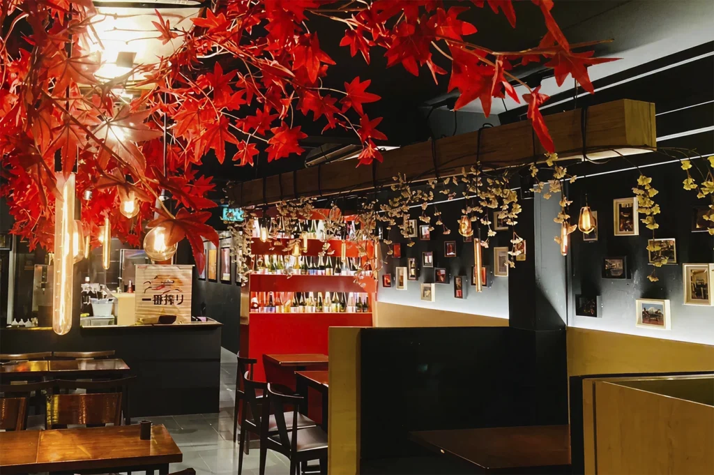 The interior of the restaurant, decorated with artificial red maple branches hanging from the ceiling and warm Edison bulb lighting, featuring wooden tables, dark seating, and a back wall displaying rows of sake bottles.