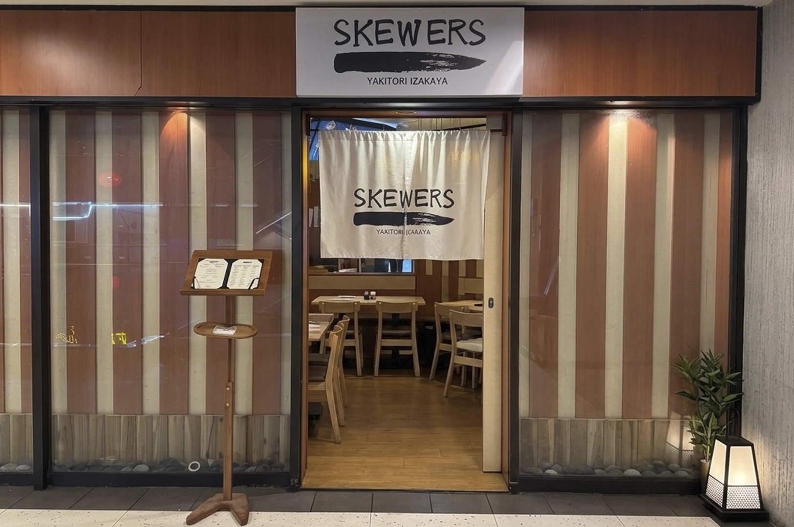 The entrance to "SKEWERS," a yakitori izakaya, with a wooden-paneled storefront, a white fabric noren curtain over the doorway, and a wooden menu stand in front.