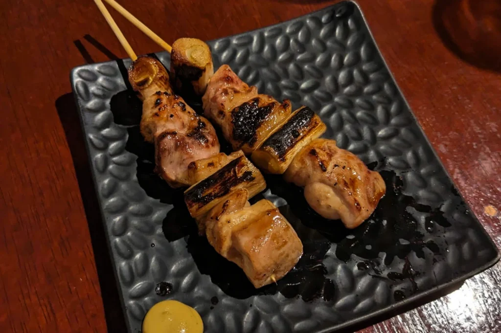 A close-up, top-down shot of two yakitori skewers resting on a textured, matte black rectangular plate. The skewers feature succulent, grilled pieces of chicken alternated with thick sections of charred leek, all glistening with a dark, savory glaze. A small dollop of yellow hot mustard sits in the bottom left corner of the plate, which is placed on a dark, rustic wooden tabletop.