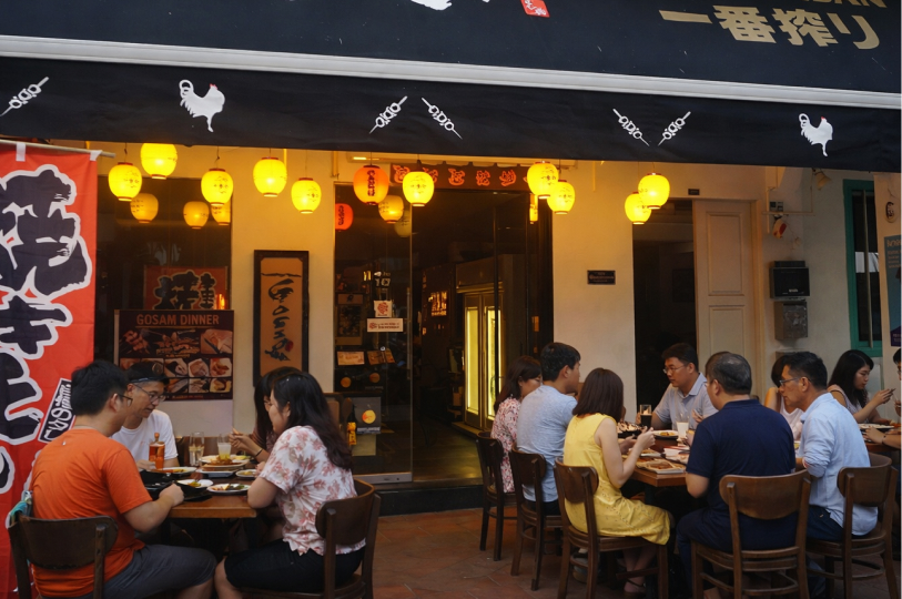 A group of people dining at outdoor tables in front of a Japanese restaurant decorated with glowing yellow lanterns.