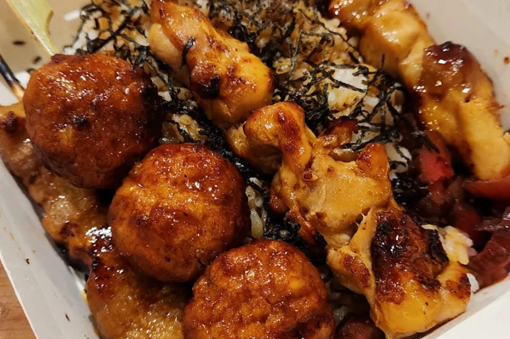 view of a yakitori bento box. The image focuses on several skewers resting on a bed of white rice topped with shredded seaweed (nori). On the left, two round, glazed chicken meatballs (tsukune) are prominent. To the right, pieces of grilled chicken thigh show charred edges and a thick, glistening savory glaze. The rice underneath is lightly stained with the dark sauce, and a small portion of reddish pickled ginger is visible in the bottom right corner. The lighting is warm and highlights the texture of the grilled meat.