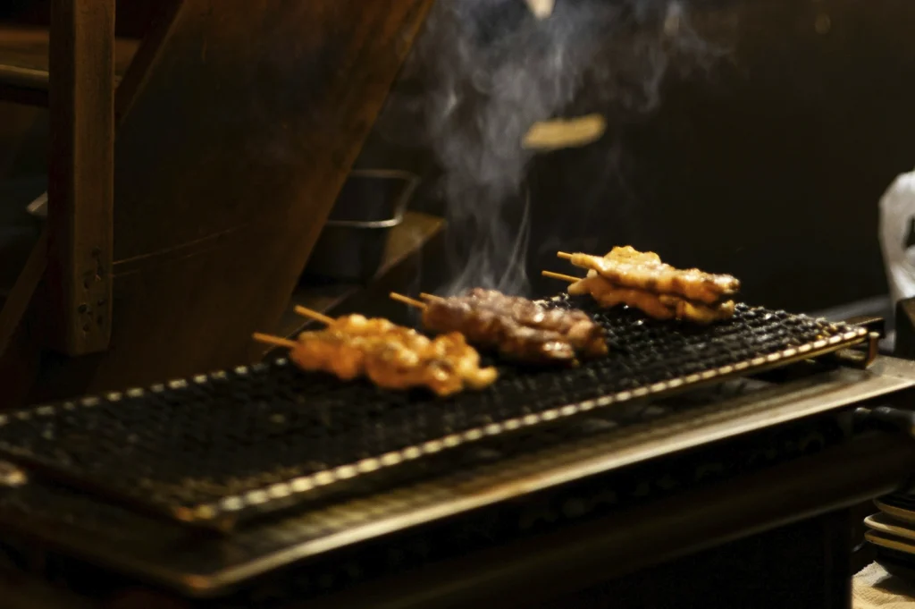 A moody, low-light photograph focusing on several yakitori skewers cooking on a narrow charcoal grill. Smoke curls elegantly upward from the meat, which is charring over the heat. The background is softly blurred, showing dark wooden architectural elements, creating a focused, atmospheric "action shot" of the grilling process.