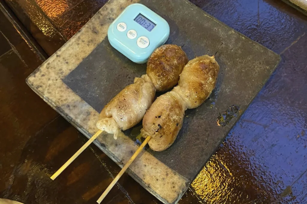 A close-up view of two grilled meat skewers served on a dark, textured slate plate, accompanied by a small, light-blue digital kitchen timer resting on the plate.