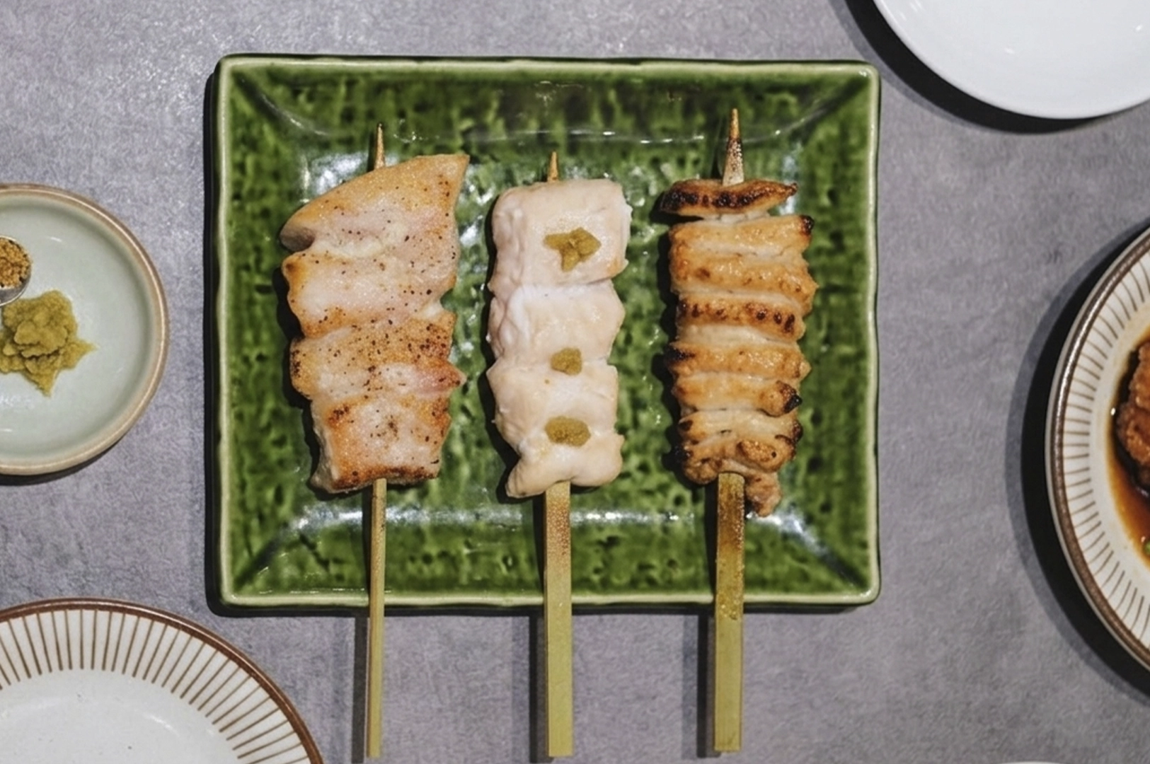 A top-down shot of three yakitori skewers—one slightly charred, one with dollops of green condiment, and one lighter in color—arranged on a green textured plate, with small dishes of seasoning on the side.