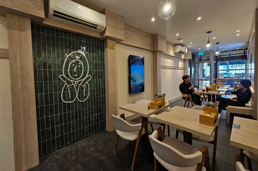 A wide-angle shot of a brightly lit, modern ramen shop with a minimalist aesthetic. The space features light wood paneling, clean white walls, and several small square tables paired with gray upholstered chairs with wooden frames. A prominent accent wall is covered in dark green vertical tiles and features a white neon-style outline of a whimsical character. In the background, two patrons are seated at different tables, and a glass storefront reveals a glimpse of the street outside.