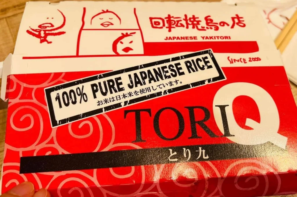 Tori Q bento box lid. The packaging is primarily red and white with a swirling pattern. The large "TORI Q" logo is printed in black and white on the lower right. A prominent white rectangular label with a black border across the center reads "100% PURE JAPANESE RICE" in bold capital letters, with Japanese text underneath. The top section of the box features red line-art illustrations of the chick mascot in various poses and the phrase "Japanese Yakitori" with "Since 2000" written in small red text.