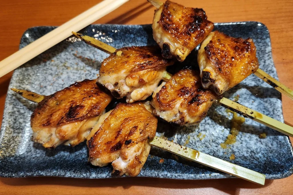 A top-down view of three skewers of grilled chicken wings (Teba) served on a blue and gray mottled rectangular plate. The wings have a crispy, deeply caramelized skin with visible charred edges. A pair of light-colored wooden chopsticks rests across the top corner of the plate on a warm-toned wooden table.