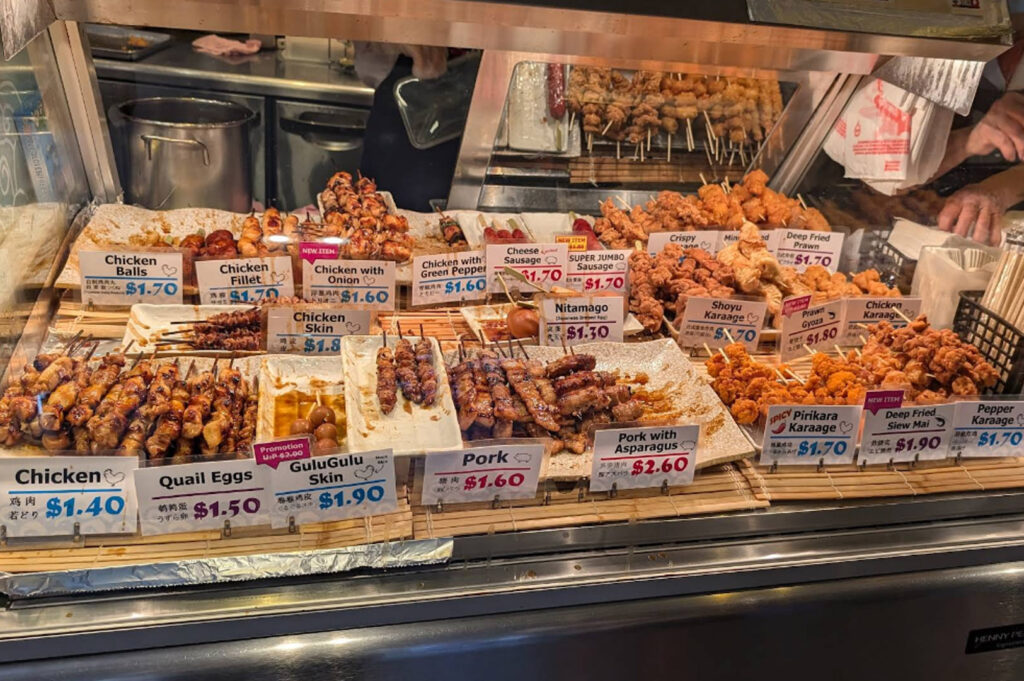 A brightly lit glass display counter at a food stall showcasing a wide variety of Japanese skewers. Each tray has a clear price tag in blue or red text. Options include "Chicken Balls" ($1.70), "GuluGulu Skin" ($1.90), "Pork with Asparagus" ($2.60), and various types of "Karaage" (fried chicken). The skewers are neatly organized on bamboo mats, and a worker's hands are visible in the upper right corner reaching for a paper bag.