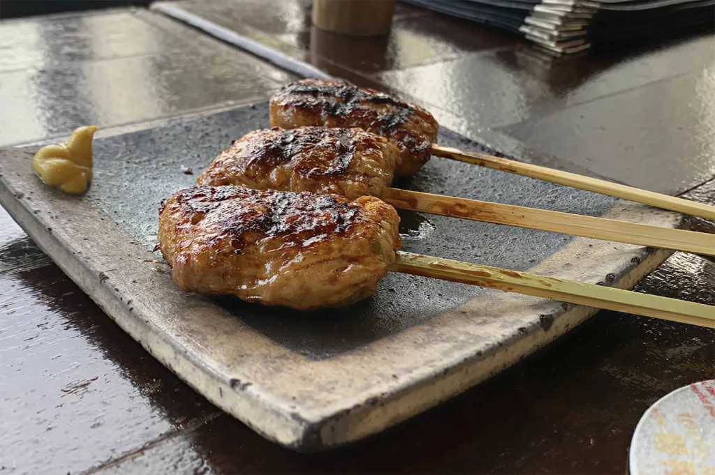 A side-angle view of three glazed, grilled skewers served on a rectangular slate plate, with a dollop of yellow mustard on the side.