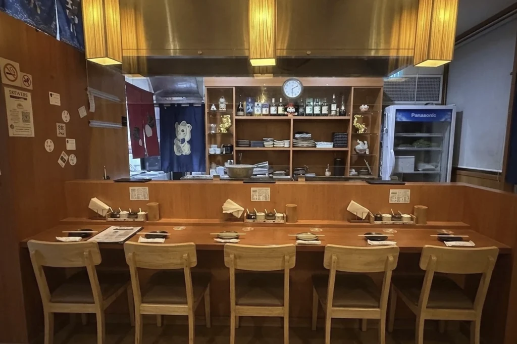 An interior view of the restaurant featuring a long wooden counter with five chairs, a well-lit kitchen prep area behind it with shelving holding bottles, and a clock on the wall.