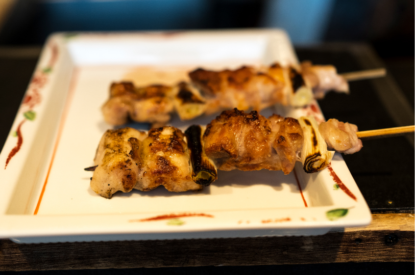 Two finished chicken and scallion yakitori skewers (negima) served on a rectangular white plate.