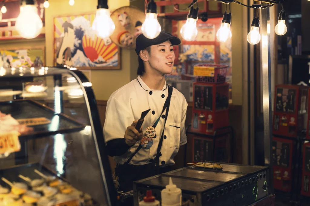 A candid shot of a young chef in a white double-breasted uniform and a black cap, working behind a Japanese food stall. He is holding a small spatula and looking off-camera. The scene is warmly lit by several hanging incandescent light bulbs, with traditional Japanese artwork and "Gachapon" capsule machines visible in the blurred background.