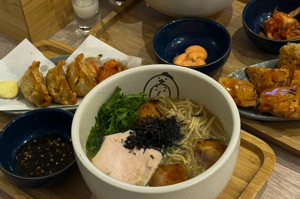 A top-down view of a wooden tray filled with various Japanese dishes, centered around a large white bowl of ramen. The ramen contains noodles in a light broth, topped with a slice of chicken, shredded nori, and greens, with a small chicken logo printed on the inside rim of the bowl. Surrounding the main bowl are side dishes including golden-brown fried gyoza, a small bowl of kimchi, a dark dipping sauce, and a plate of glazed grilled chicken pieces, all presented in a clean, appetizing manner.