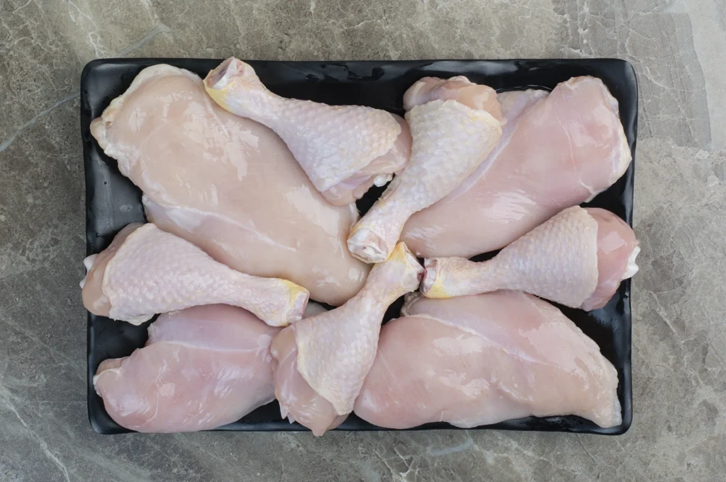 A clean, overhead studio shot of raw chicken parts arranged on a rectangular black tray. The tray contains two large chicken breasts and several drumsticks with the skin on. The meat is pale pink and fresh, set against a neutral, light gray marbled stone surface.