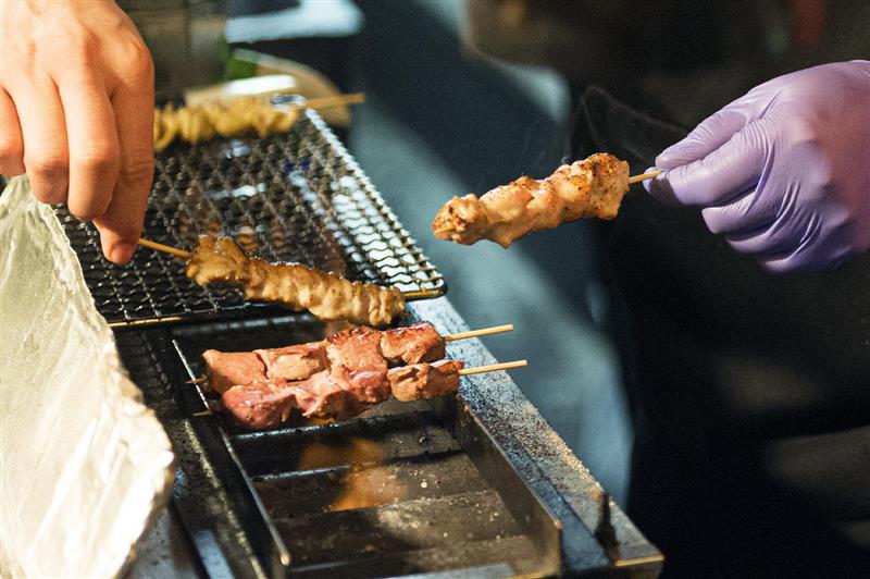 A close-up, action shot of yakitori being grilled. On the left, a bare hand uses a skewer to adjust meat on a wire mesh rack over glowing coals. On the right, a hand wearing a purple nitrile glove holds up a perfectly grilled, seasoned chicken skewer. The background is dark and blurry, highlighting the textures of the charred meat and the orange glow of the fire beneath the grill.