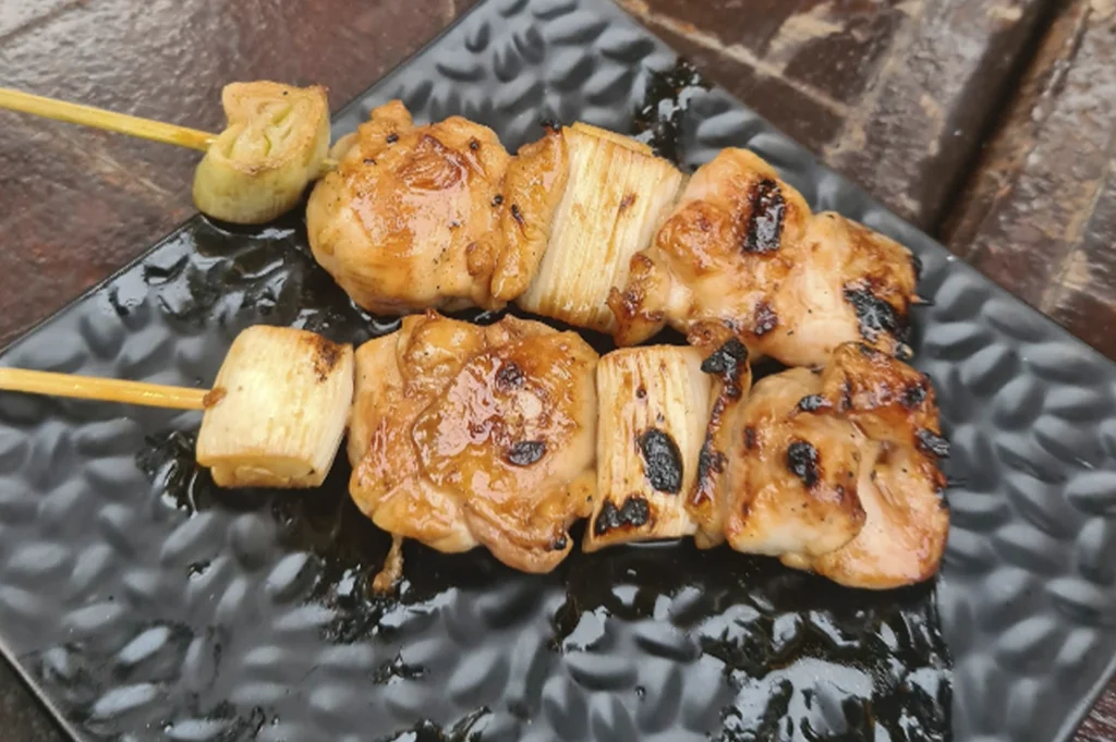 Two grilled yakitori skewers consisting of alternating pieces of chicken and leeks, served on a textured, matte black plate.