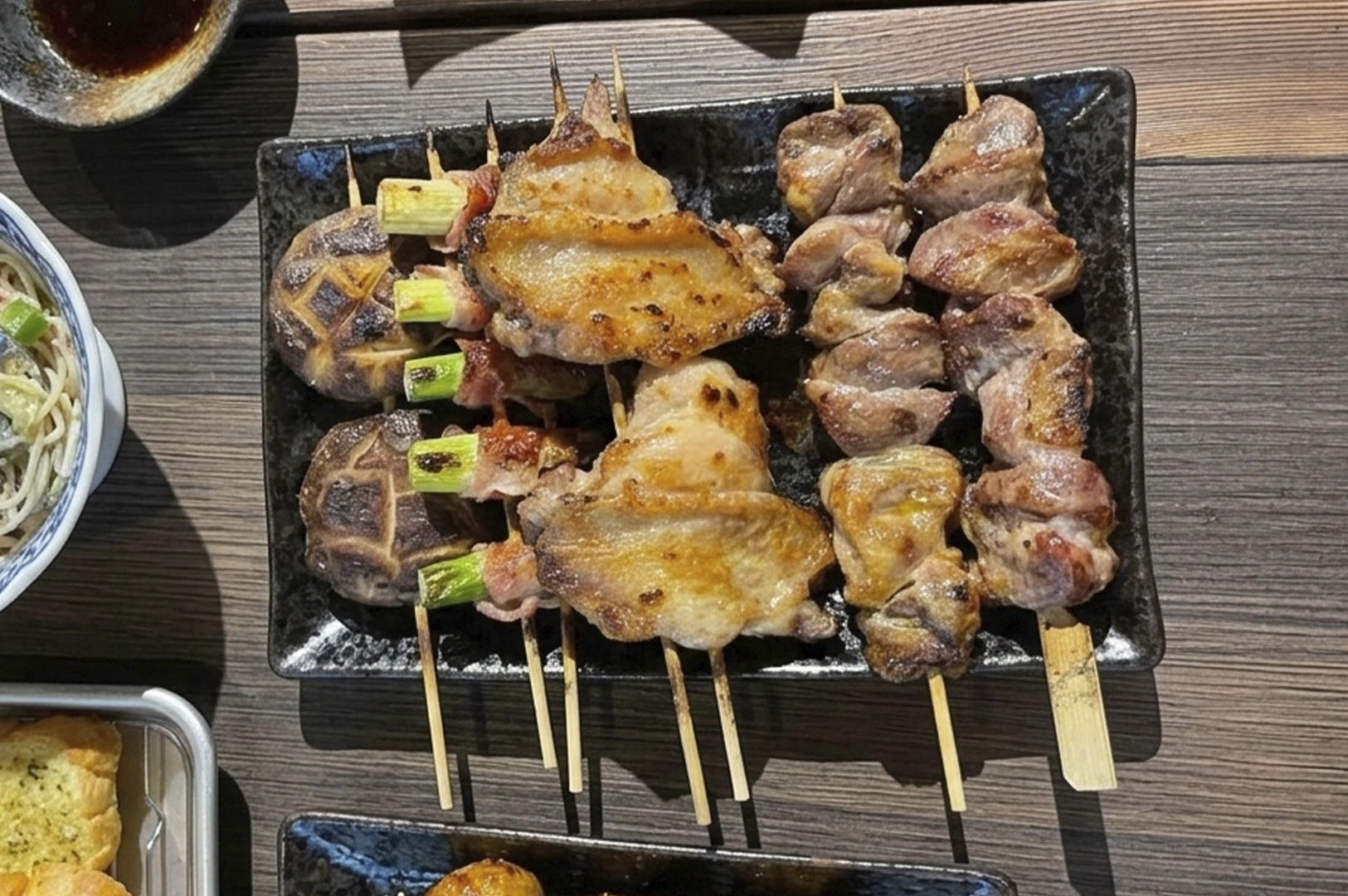 A top-down view of a rectangular black plate piled with various yakitori skewers, including grilled mushrooms, bacon-wrapped asparagus, chicken wings, and skewers of succulent meat.
