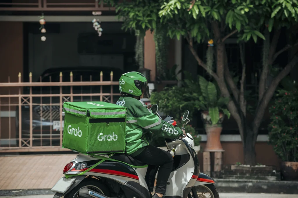 A side-profile shot of a Grab delivery rider on a white and red motor scooter, driving down a suburban street. The rider is wearing a full green Grab uniform and a matching green helmet. A large, square, green thermal delivery box with the "Grab" logo is securely strapped to the back of the scooter.