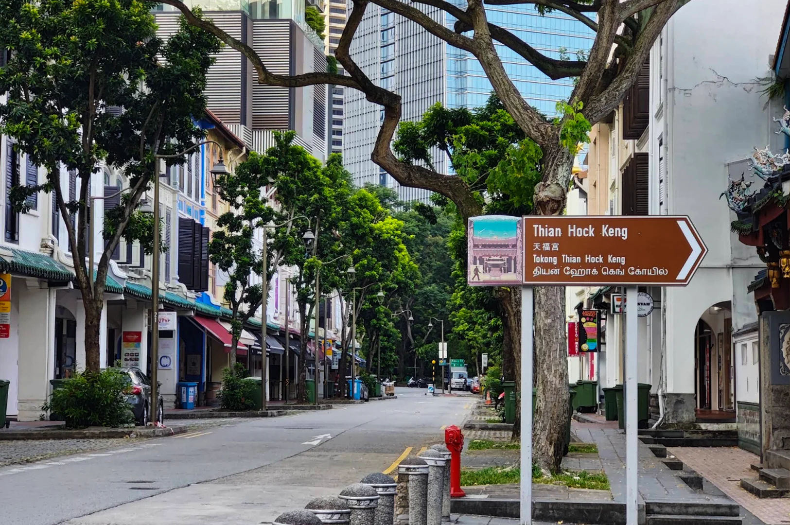 A street-level view looking down a clean, paved road in Singapore, flanked by historic two and three-story colorful shophouses with traditional shutters. In the foreground, a large brown directional sign points to the right toward "Thian Hock Keng" temple, with text written in English, Chinese, Malay, and Tamil. Lush green trees line the sidewalk, and modern glass skyscrapers rise in the background, creating a sharp contrast between heritage architecture and the contemporary city skyline.