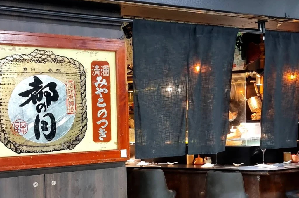 An exterior-facing view of a traditional Japanese eatery. On the left, a large framed vintage-style sign features a sake barrel illustration with bold Japanese calligraphy. Dark blue "noren" (split curtains) hang across the entrance, partially obscuring the interior where a chef can be seen working. Two dark, modern bar stools are positioned in front of a wooden counter.