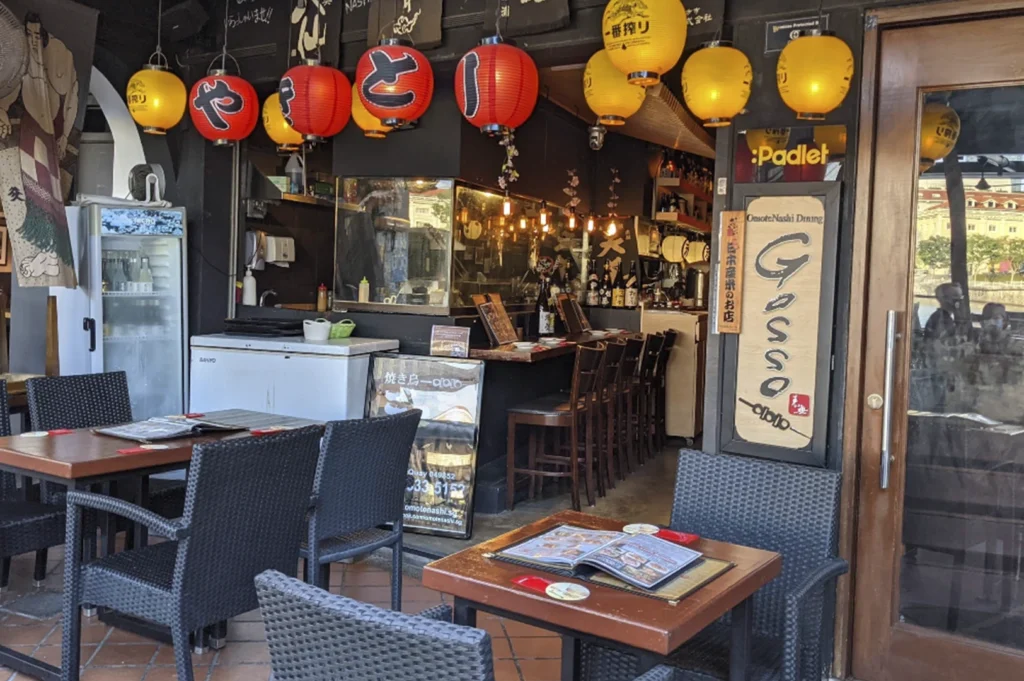 A view of the restaurant's entrance with outdoor seating, decorated with traditional Japanese red and yellow lanterns, with a sign on the door identifying it as "OmoteNashi Dining Gosso."