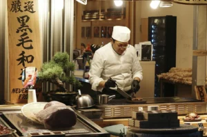 A professional chef in a white uniform, white hat, and black gloves stands behind a stainless steel counter in a Japanese restaurant. He is focused on using a spatula to flip pieces of meat on a flat-top grill. In the foreground, a large raw cut of marbled beef sits in a wicker basket on a glass display case. To the left is a wooden sign with Japanese calligraphy and a small bonsai tree.