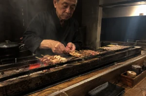 A high-angle, medium shot captures an elderly Japanese chef with a focused expression as he meticulously grills several rows of yakitori skewers over a traditional narrow charcoal grill. Dressed in a dark navy samue, he uses both hands to adjust the wooden skewers, which feature various cuts of chicken and green onions charred to a golden brown. The grill glows with intense orange embers from binchotan charcoal, sending thin wisps of white smoke into the dimly lit, atmospheric restaurant. In the foreground, a rustic wooden counter holds small ceramic condiment containers and a black rectangular serving plate, while the dark, blurred background highlights the chef’s craftsmanship and the warmth of the hearth.