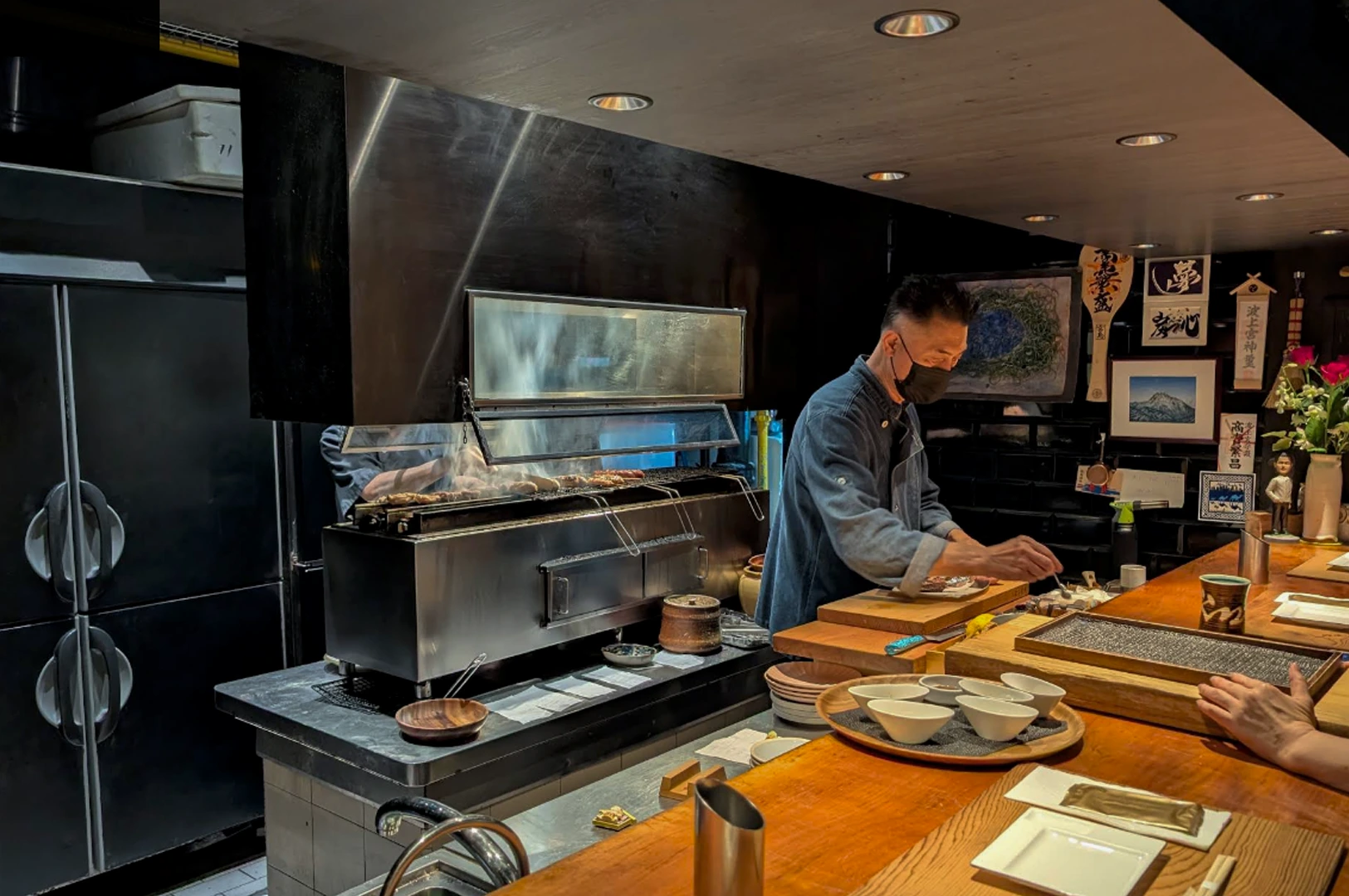 A chef working behind a wooden counter at Kazu Sumiyaki. The chef, wearing a blue denim shirt and a black face mask, is carefully plating food. To his left, a large stainless steel charcoal grill (sumiyaki) is topped with skewers of meat, with visible smoke being drawn into a large black ventilation hood above. The background features dark walls decorated with Japanese calligraphy, small framed pictures, and a small flower arrangement.